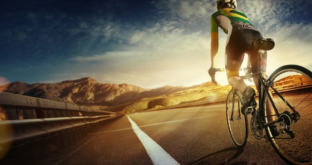 A cyclist in professional gear rides on an open road, surrounded by a mountain landscape. Like a vet devoted to their craft, the biker navigates the terrain with precision as the sun sets, casting a warm glow. A clear sky with a few clouds completes this picturesque scene.