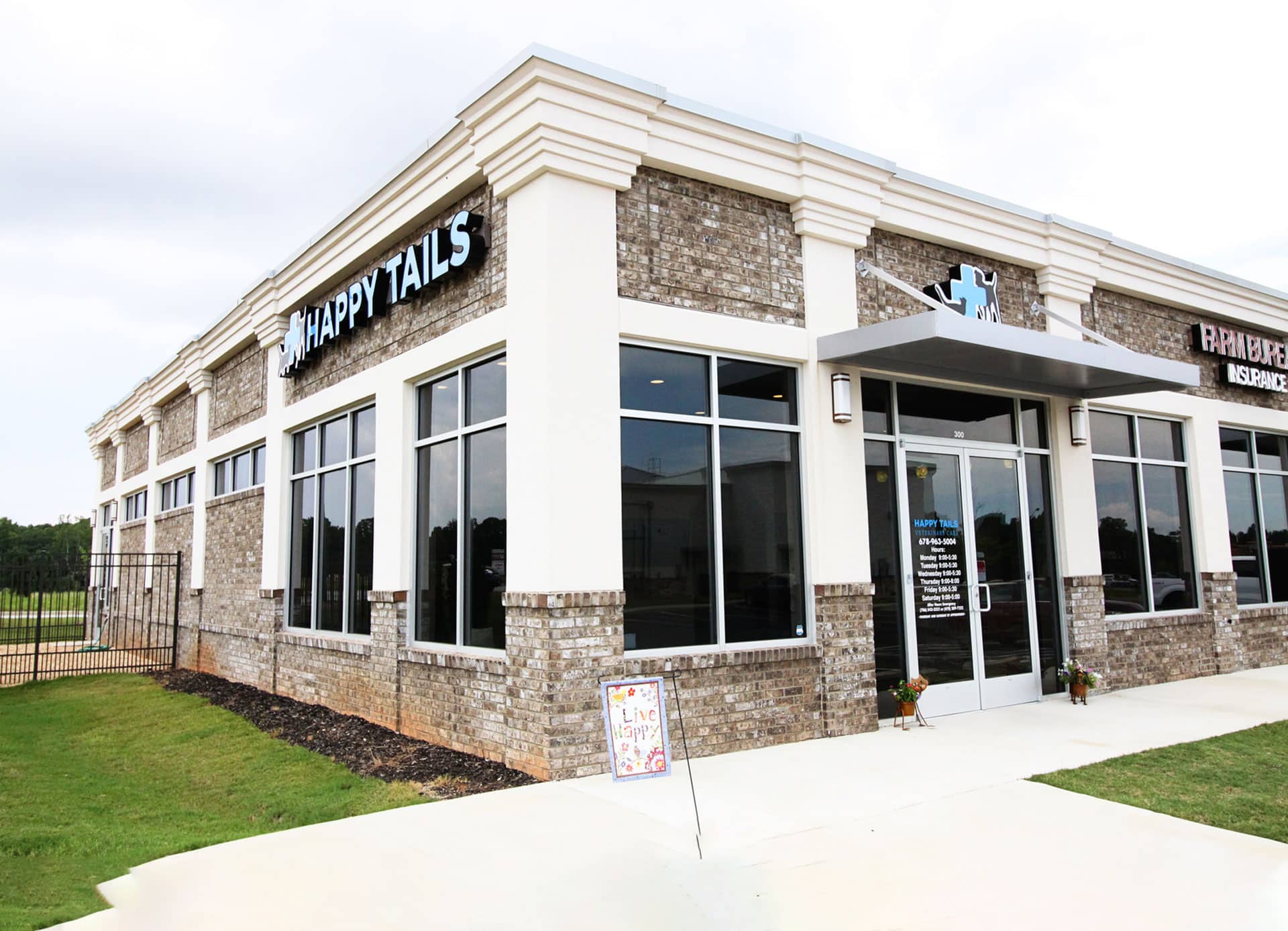 A corner view of a modern building with large windows and brick detailing houses "Happy Tails Vet." A sidewalk leads to the entrance, where a small decorative sign is placed outside. The sky is overcast, adding a somber tone to this bustling veterinarian clinic.