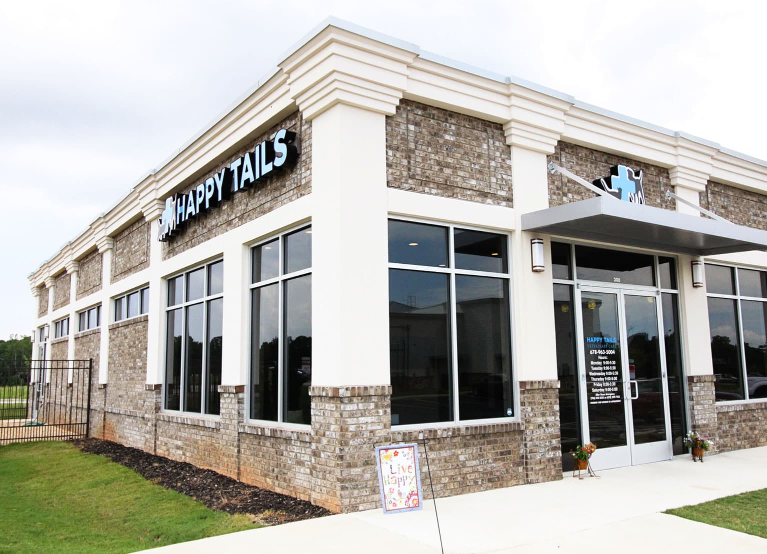 A modern brick and glass building with a sign reading "Happy Tails Veterinary." The entrance features a door with business hours posted. A small signboard with colorful lettering is placed near the entrance, indicating it's a trusted vet clinic. The sky is overcast.